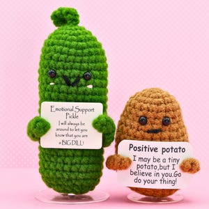 May include: Two crocheted plushies, a green pickle and a brown potato, holding signs with encouraging messages. The pickle's sign reads "Emotional Support Pickle I will always be around to let you know that you are a BIG DILL!" The potato's sign reads "Positive potato I may be a tiny potato, but I believe in you. Go do your thing!"
