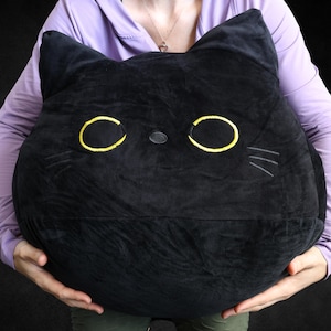 May include: A large black plush cat pillow with yellow eyes and a black nose. The pillow is being held by a person's hands.
