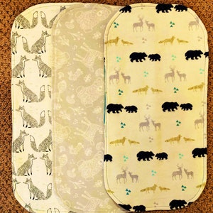 May include: Three fabric burp cloths with different patterns. The first cloth has a white background with a black fox pattern. The second cloth has a white background with a brown bear and deer pattern. The third cloth has a white background with a black bear and deer pattern.