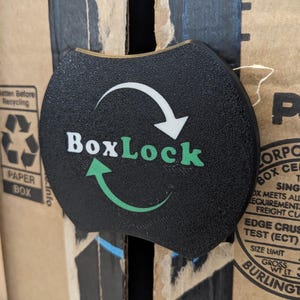 May include: A black and green circular sign with a white arrow pointing clockwise and the text "BoxLock" in white letters. The sign is attached to a cardboard box.