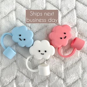 May include: Three cloud-shaped bottle stoppers in pastel colors: light blue, white, and pink. Each stopper has a smiling face and a matching colored loop and cap. The stoppers are designed to fit bottles.