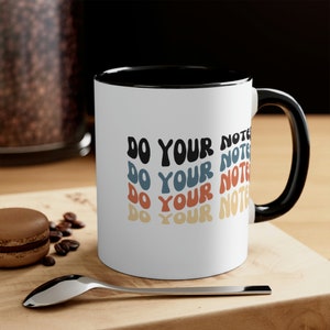May include: White ceramic coffee mug with a black rim and handle. The mug has the text "DO YOUR NOTES" repeated four times in a retro font with different colored backgrounds.