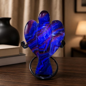 May include: A polished stone carving in the shape of an angel, featuring vibrant bands of deep blue and purple hues. The figure is displayed upright on a black metal stand, showcasing a smooth, glossy surface with natural mineral patterns.