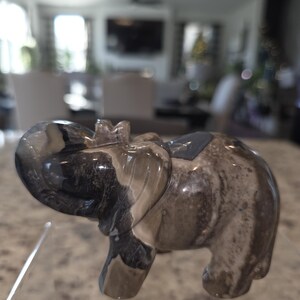 May include: A polished stone elephant figurine featuring a marbled pattern of dark charcoal, grey, and beige tones. The carving depicts an elephant with its trunk curled upward, showcasing smooth, glossy surfaces and natural mineral veining throughout.