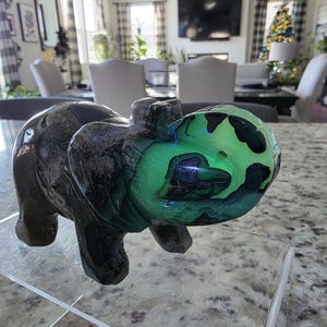 May include: A carved stone elephant figurine featuring a dark brown and black body with a vibrant, glowing neon green patch on its head and trunk. The text "Michelle Crystals" is printed in white at the bottom left of the image.