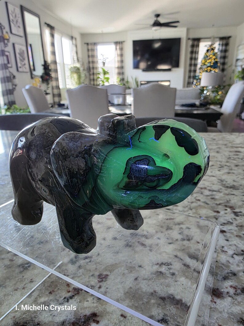 May include: A carved stone elephant figurine featuring a dark brown and black body with a vibrant, glowing neon green patch on its head and trunk. The text "Michelle Crystals" is printed in white at the bottom left of the image.