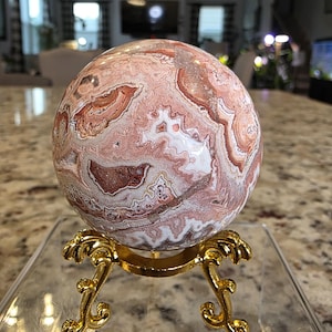 May include: A polished spherical stone featuring intricate swirling patterns in shades of terracotta, cream, and white, resting on a gold-toned metal tripod stand. The text "The Elevated Stone" is printed in white at the bottom of the image.