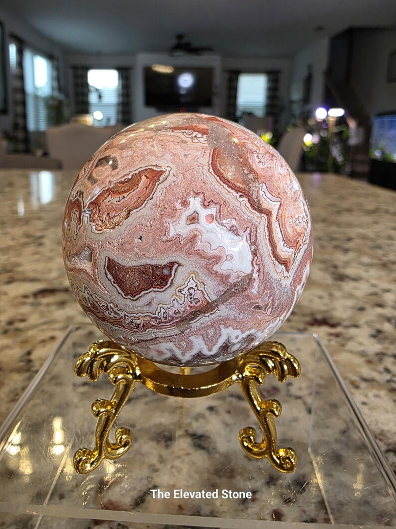 May include: A polished spherical stone featuring intricate swirling patterns in shades of terracotta, cream, and white, resting on a gold-toned metal tripod stand. The text "The Elevated Stone" is printed in white at the bottom of the image.