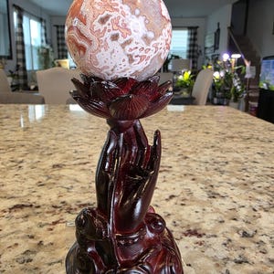 May include: A polished, spherical stone with intricate white, tan, and reddish-brown patterns resting on a dark reddish-brown resin stand shaped like a lotus flower held by a hand. The base features the text The Elevated Stone at the bottom.