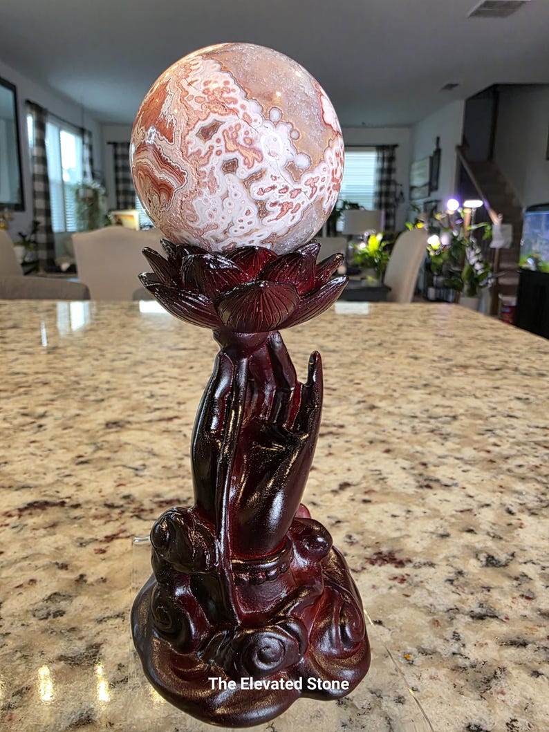 May include: A polished, spherical stone with intricate white, tan, and reddish-brown patterns resting on a dark reddish-brown resin stand shaped like a lotus flower held by a hand. The base features the text The Elevated Stone at the bottom.
