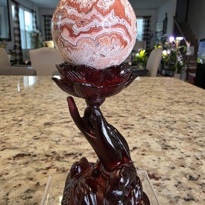 May include: A polished, spherical stone with intricate, swirling patterns in shades of reddish-brown, cream, and white. It rests upon a dark, reddish-brown stand shaped like a hand holding a lotus flower, with the text The Elevated Stone at the base.