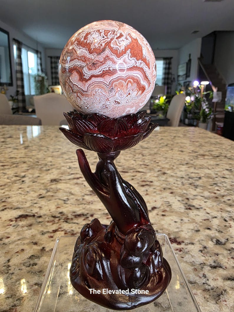 May include: A polished, spherical stone with intricate, swirling patterns in shades of reddish-brown, cream, and white. It rests upon a dark, reddish-brown stand shaped like a hand holding a lotus flower, with the text The Elevated Stone at the base.