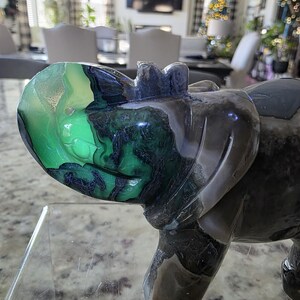 May include: A polished stone carving of a rhinoceros featuring a vibrant green and black patterned head and a dark grey body. The sculpture displays natural mineral banding and color variations throughout its surface. The text I. Michelle Crystals is visible in the bottom left corner.