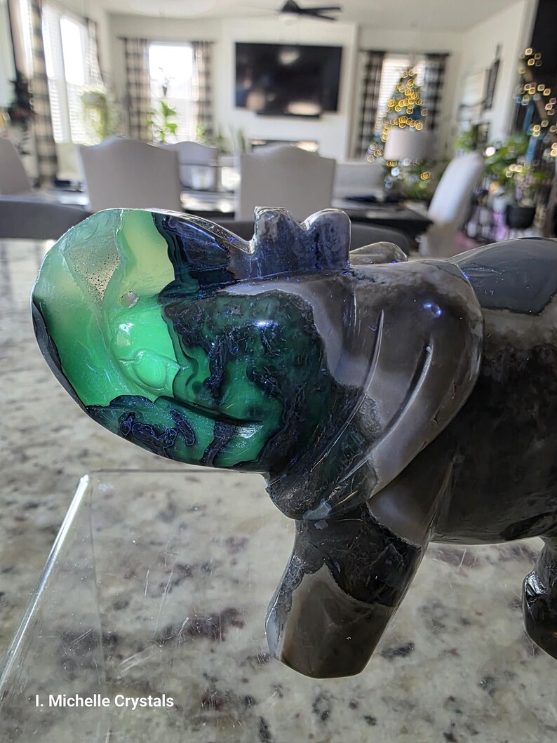 May include: A polished stone carving of a rhinoceros featuring a vibrant green and black patterned head and a dark grey body. The sculpture displays natural mineral banding and color variations throughout its surface. The text I. Michelle Crystals is visible in the bottom left corner.