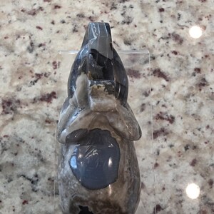 May include: A carved stone elephant figurine featuring a polished surface with mottled gray, brown, and white patterns. The sculpture displays a smooth texture with a prominent dark gray circular patch on the torso and a clear acrylic stand.