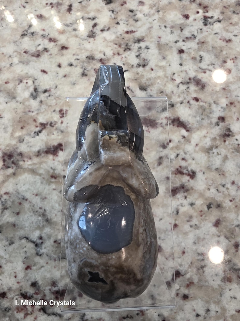 May include: A carved stone elephant figurine featuring a polished surface with mottled gray, brown, and white patterns. The sculpture displays a smooth texture with a prominent dark gray circular patch on the torso and a clear acrylic stand.
