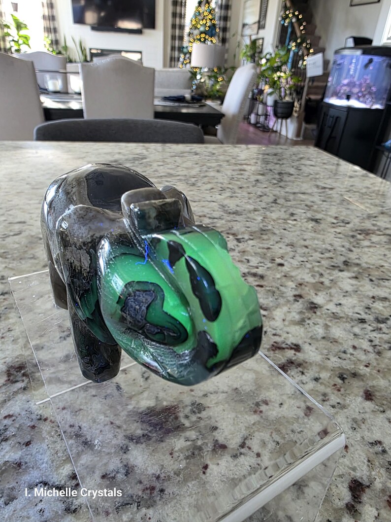 May include: A carved stone elephant figurine featuring a polished surface with deep black and vibrant green marbled patterns. The elephant is positioned on a clear acrylic stand, and the text "I. Michelle Crystals" is visible in the bottom left corner.