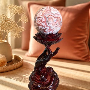 May include: A spherical stone with intricate pink, white, and grey swirling patterns resting atop a dark red lotus flower held by a sculpted hand base. The object features a polished finish and sits vertically as a decorative display piece.