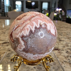 May include: A spherical stone with banded patterns in shades of pink, white, and brown, featuring a central geode cavity with clear crystalline structures. The stone rests on a gold-colored metal stand with ornate, curved legs and feet.