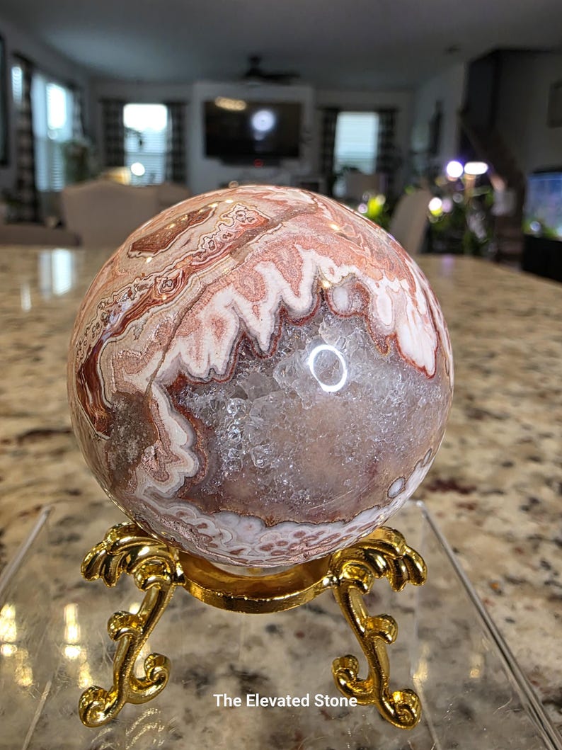 May include: A spherical stone with banded patterns in shades of pink, white, and brown, featuring a central geode cavity with clear crystalline structures. The stone rests on a gold-colored metal stand with ornate, curved legs and feet.