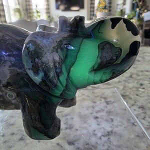 May include: A polished stone carving of an elephant featuring vibrant green, black, and grey swirling patterns throughout its body. The sculpture has a smooth, glossy finish and is displayed on a clear acrylic stand. Text reads I. Michelle Crystals.