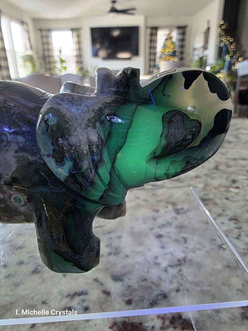 May include: A polished stone carving of an elephant featuring vibrant green, black, and grey swirling patterns throughout its body. The sculpture has a smooth, glossy finish and is displayed on a clear acrylic stand. Text reads I. Michelle Crystals.