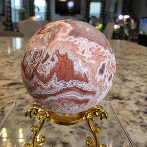 May include: A polished spherical stone featuring intricate swirling patterns in shades of rust, cream, and white. The sphere rests on a gold-colored metal stand with ornate, curved legs. The text "The Elevated Stone" is printed at the bottom.