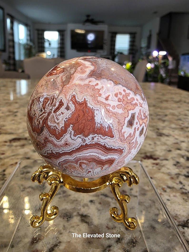 May include: A polished spherical stone featuring intricate swirling patterns in shades of rust, cream, and white. The sphere rests on a gold-colored metal stand with ornate, curved legs. The text "The Elevated Stone" is printed at the bottom.