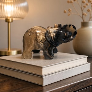 May include: A carved stone elephant figurine featuring a smooth, polished surface with a color gradient transitioning from a light beige, speckled body to a deep, solid black head and trunk, standing in a profile position with its trunk curled upward.