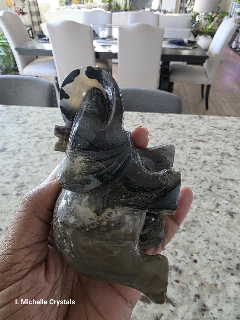 May include: A carved stone elephant figurine featuring a polished surface with swirling patterns in shades of black, gray, and translucent white. The sculpture displays natural banding and mineral inclusions throughout its form. Text reads I. Michelle Crystals.