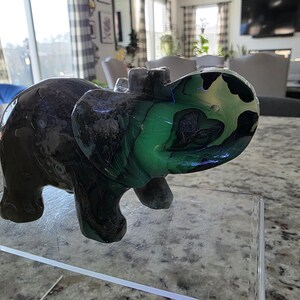 May include: A carved stone elephant figurine featuring a dark brown body with a vibrant, translucent green and black marbled pattern on the head and trunk. The text "I. Michelle Crystals" is printed in white at the bottom left of the image.