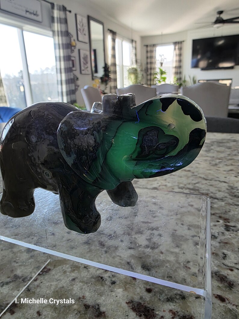 May include: A carved stone elephant figurine featuring a dark brown body with a vibrant, translucent green and black marbled pattern on the head and trunk. The text "I. Michelle Crystals" is printed in white at the bottom left of the image.