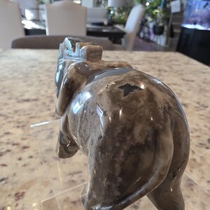 May include: A polished stone carving of an elephant in shades of gray, brown, and white with a smooth, glossy finish. The figurine is depicted from a rear-facing angle, showing its rounded back, tail, and ears. Text reads I. Michelle Crystals.