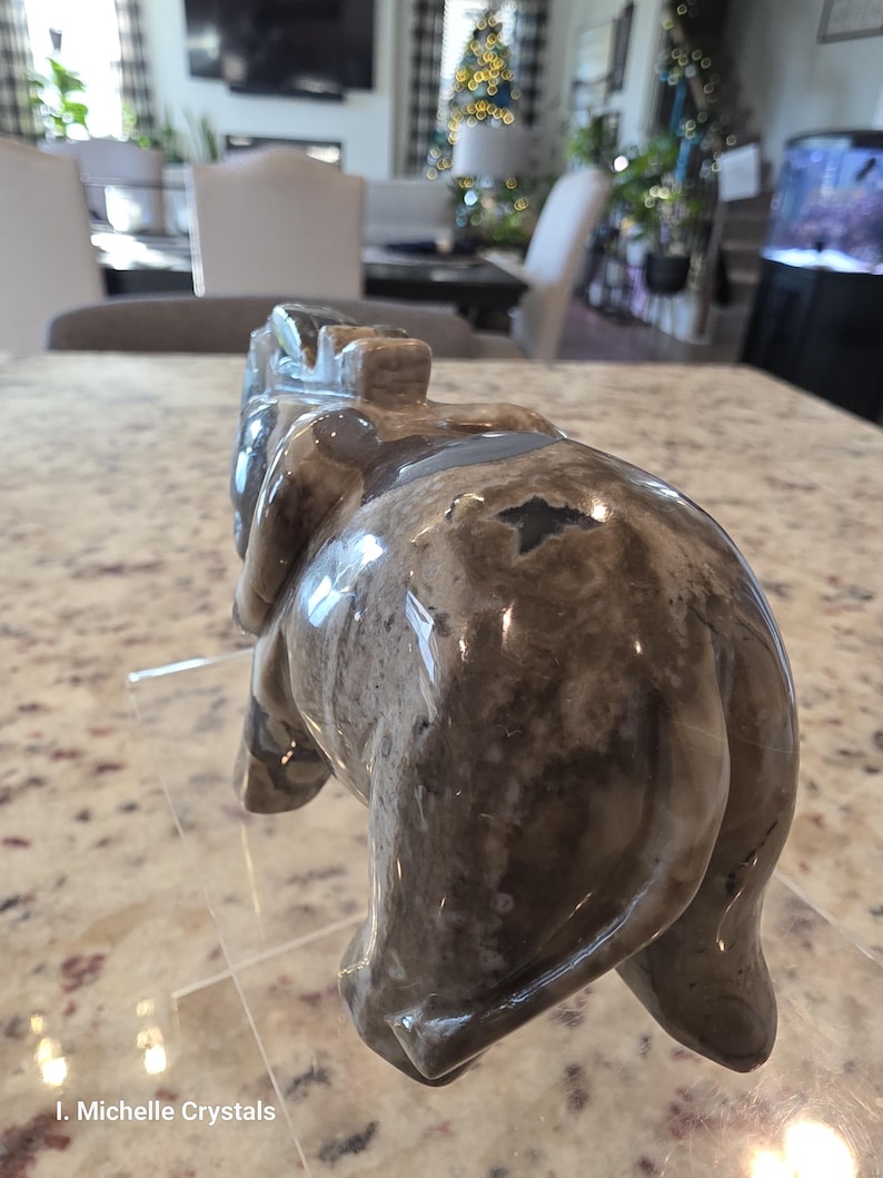 May include: A polished stone carving of an elephant in shades of gray, brown, and white with a smooth, glossy finish. The figurine is depicted from a rear-facing angle, showing its rounded back, tail, and ears. Text reads I. Michelle Crystals.