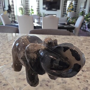 May include: A polished stone elephant figurine featuring a dark gray and black marbled pattern with smooth, rounded contours. The carving displays a trunk curled upward and visible ear details. The text I. Michelle Crystals is printed at the bottom.