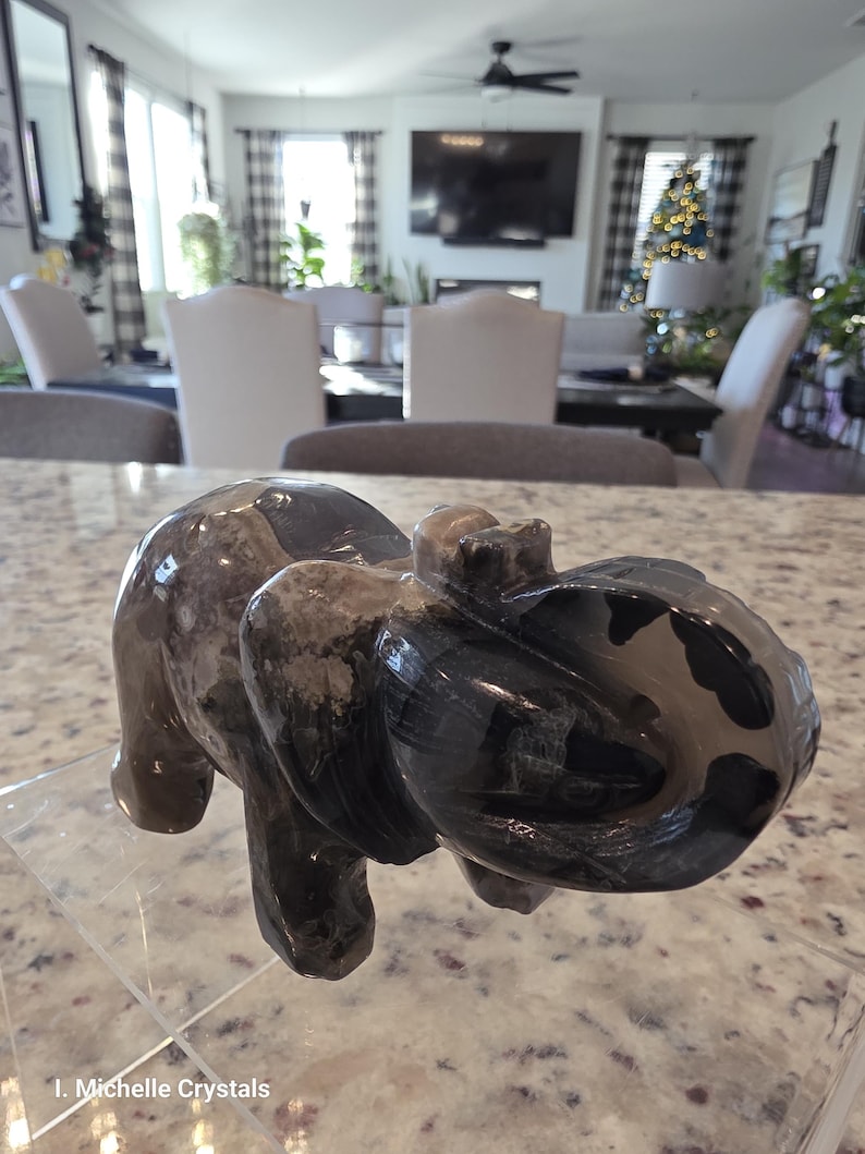 May include: A polished stone elephant figurine featuring a dark gray and black marbled pattern with smooth, rounded contours. The carving displays a trunk curled upward and visible ear details. The text I. Michelle Crystals is printed at the bottom.