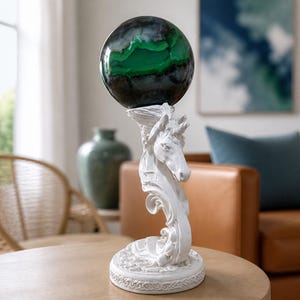 May include: A white unicorn-shaped stand with a circular base supporting a large, polished spherical stone. The stone features deep black and dark green banded patterns with a smooth, glossy surface. The stand is sculpted with intricate details.