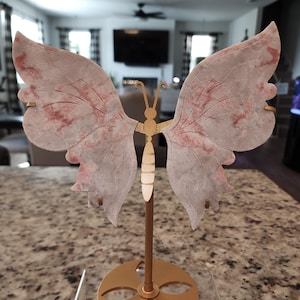 May include: A decorative butterfly sculpture with a gold-colored metal body and stand. The butterfly wings are a translucent pink with red veining. The sculpture is displayed on a speckled countertop.