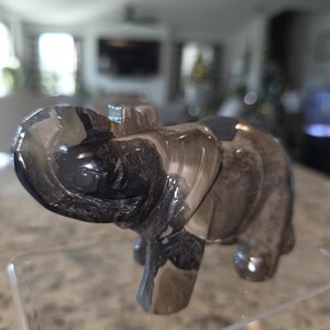 May include: A carved elephant figurine made of polished smoky quartz with translucent grey and black banded patterns. The elephant is depicted with its trunk raised upward and is positioned on a clear acrylic stand. The text I. Michelle Crystals is visible.