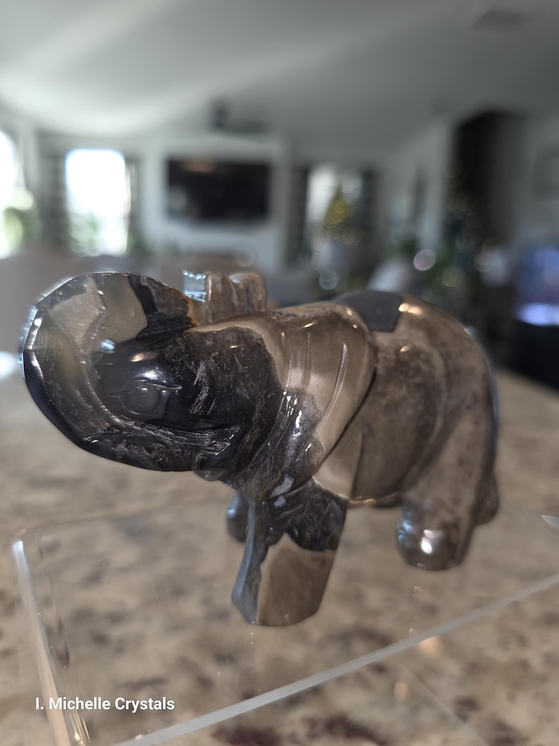 May include: A carved elephant figurine made of polished smoky quartz with translucent grey and black banded patterns. The elephant is depicted with its trunk raised upward and is positioned on a clear acrylic stand. The text I. Michelle Crystals is visible.