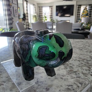 May include: A carved stone elephant figurine featuring a polished surface with deep black and vibrant neon green color patterns. The sculpture displays a smooth, glossy texture throughout its form, with the text I. Michelle Crystals visible.