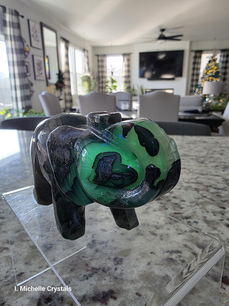 May include: A carved stone elephant figurine featuring a polished surface with deep black and vibrant neon green color patterns. The sculpture displays a smooth, glossy texture throughout its form, with the text I. Michelle Crystals visible.