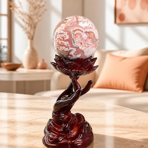 May include: A polished, spherical stone with swirling patterns of pink, white, and brown resting atop a dark reddish-brown resin sculpture. The sculpture depicts a pair of hands held upward in a cupped position, forming a lotus flower shape.