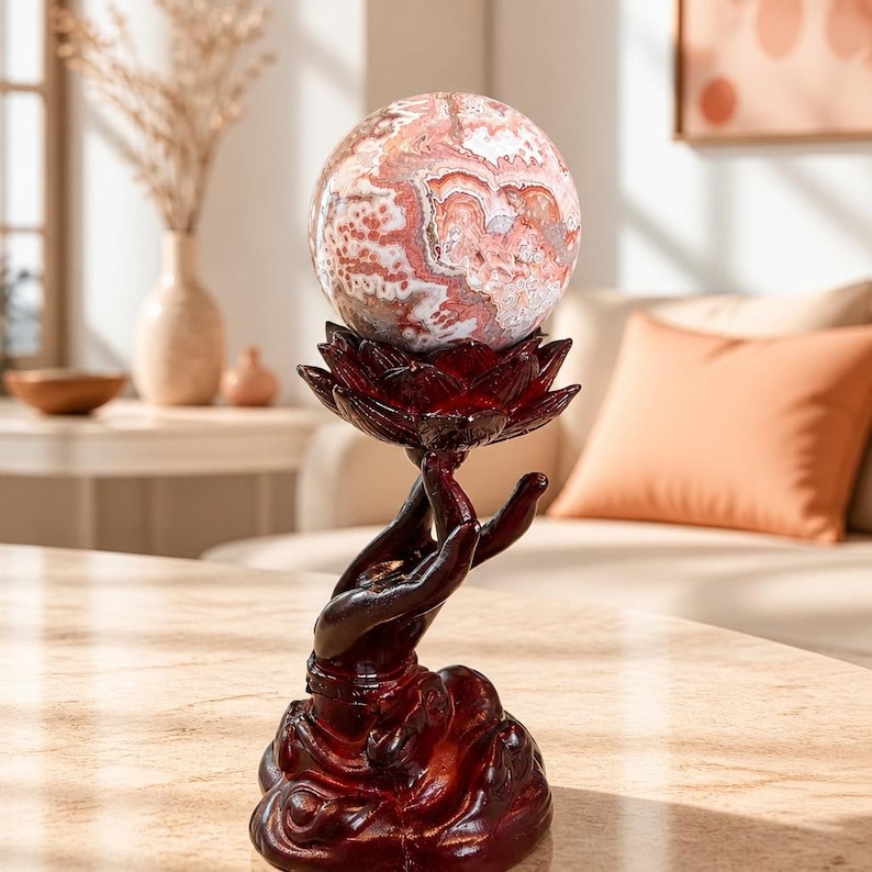 May include: A polished, spherical stone with swirling patterns of pink, white, and brown resting atop a dark reddish-brown resin sculpture. The sculpture depicts a pair of hands held upward in a cupped position, forming a lotus flower shape.