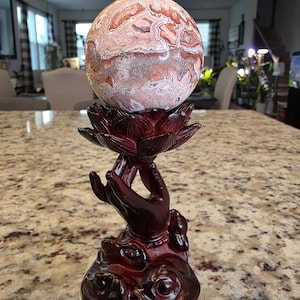May include: A spherical stone with intricate swirling patterns in shades of cream, tan, and reddish-brown, resting atop a dark reddish-brown resin stand shaped like a hand holding a lotus flower. The text "The Elevated Stone" is visible at the base.