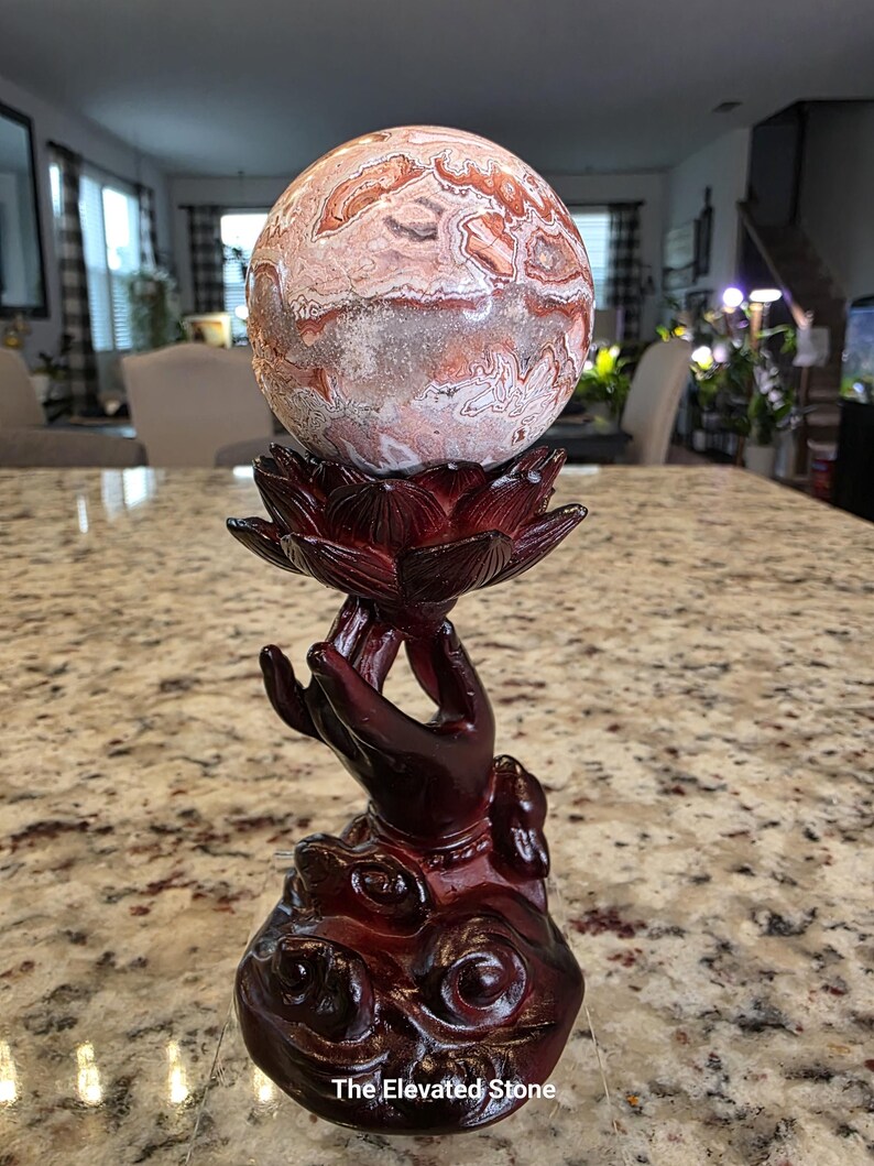 May include: A spherical stone with intricate swirling patterns in shades of cream, tan, and reddish-brown, resting atop a dark reddish-brown resin stand shaped like a hand holding a lotus flower. The text "The Elevated Stone" is visible at the base.