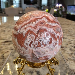 May include: A polished spherical stone featuring intricate, swirling bands of reddish-brown, cream, and white mineral patterns. The sphere rests on a decorative, ornate gold-colored metal tripod stand. The text The Elevated Stone is visible.
