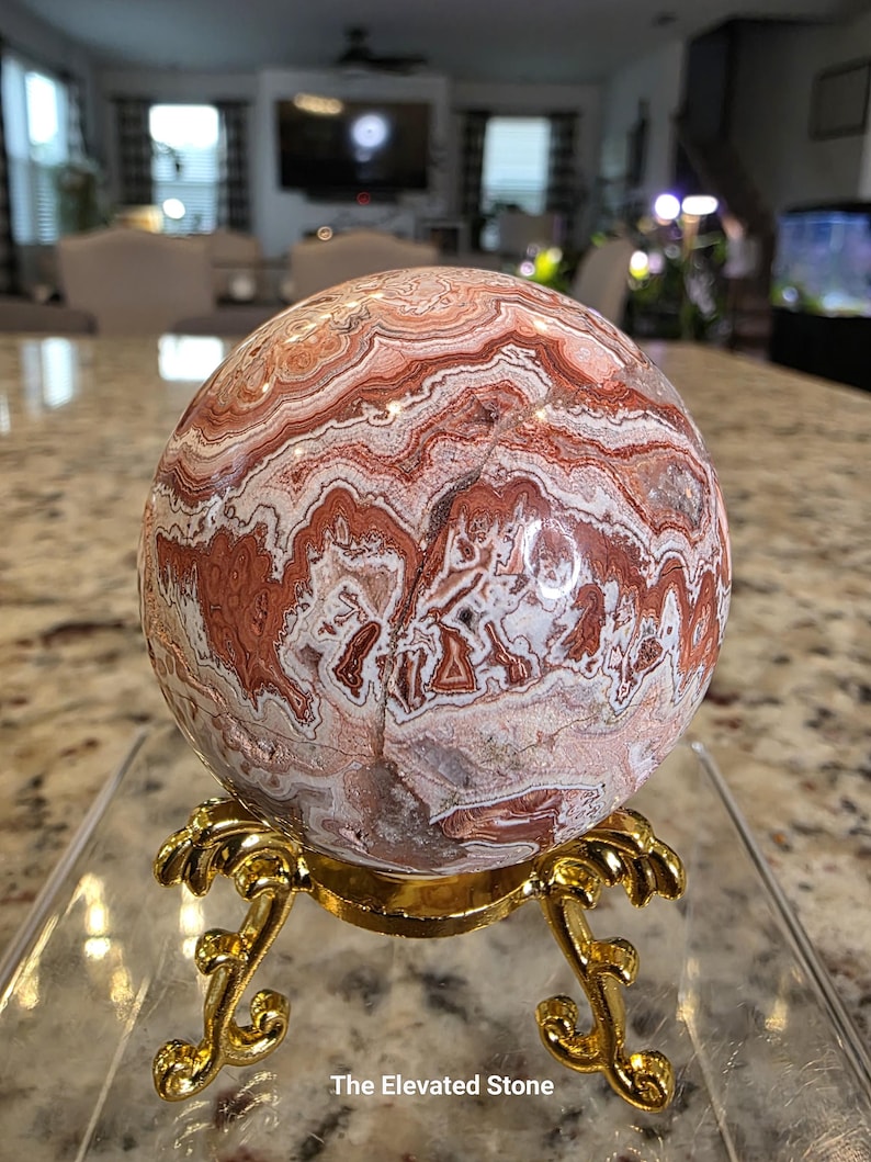May include: A polished spherical stone featuring intricate, swirling bands of reddish-brown, cream, and white mineral patterns. The sphere rests on a decorative, ornate gold-colored metal tripod stand. The text The Elevated Stone is visible.