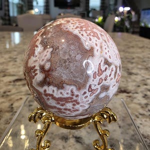May include: A spherical stone with a complex pattern of white, tan, and reddish-brown circular markings and a central crystalline geode cavity, resting on a decorative gold-colored metal tripod stand with ornate, curved legs and feet.