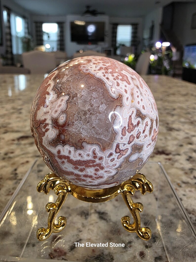 May include: A spherical stone with a complex pattern of white, tan, and reddish-brown circular markings and a central crystalline geode cavity, resting on a decorative gold-colored metal tripod stand with ornate, curved legs and feet.